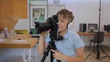 Açık mavi gömlekli genç bir çocuk parlak ışıklı bir sınıfta fotoğraf ve video çalışıyor, kulaklık takıyor ve kameranın merceğine odaklanmış ve heyecanlı bir şekilde bakıyor..