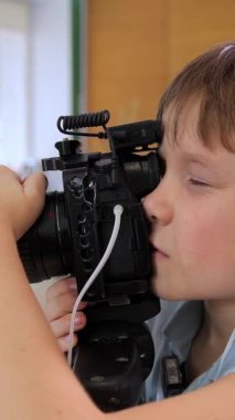 Dikey video. Kulaklık takan bir çocuk kamera tutarken dikkatle odaklanır. Parlak kapalı alan yaratıcılık ve merakın bir karışımını sergiliyor. Genç kameraman.
