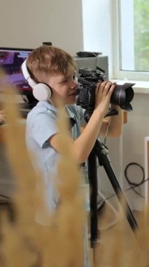Dikey video. Açık mavi gömlekli genç bir çocuk, parlak ışıklı bir odada video çalışıyor, kulaklık takıyor ve konsantrasyon ve heyecanla kameraya bakıyor..
