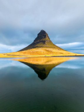 İzlanda 'daki Kirkjufell Dağı' nın solunda küçük bir gökkuşağı olan bulutlu bir günde gölde yansıması var.
