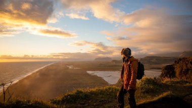İzlanda 'daki bir dağın tepesinde gün batımında denize bakan Latin bir adam.