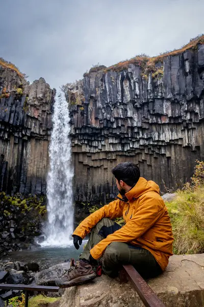 Genç Latin adam kayalıklarda oturmuş şelaleye bakıyor.