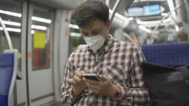 Maskeli adam Münih 'te metroya biniyor ve akıllı telefonuyla internette sörf yapıyor. Hijyen, sağlık ve enfeksiyon konsepti. Yolcu metroda seyahat ederken Coronavirus haberlerini aramak için telefonu kullanıyor. 