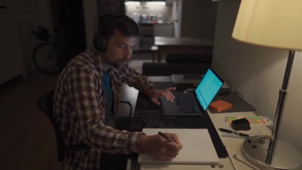ノートパソコンをヘッドフォンで勉強し 夕方にはペンでノートパソコンにメモを取る男性学生 男は夜遅くにコンピュータで働いてるアパートのヘッドセットで — ストック動画