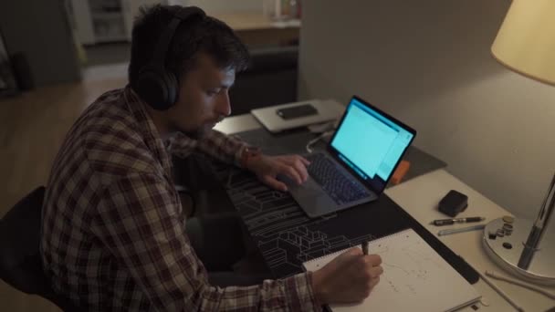 一名男生晚上带着耳机在笔记本电脑上学习 并用钢笔在笔记本上记笔记 男人深夜在电脑前工作 房间里有耳机 — 图库视频影像