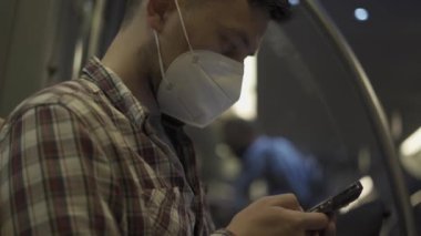 Maskeli beyaz adam Münih metrosunda sırtı pencereye dönük otururken akıllı telefon kullanıyor. Almanya 'da metroda seyahat ederken internette maske takan bir erkek. Toplu taşıma. 