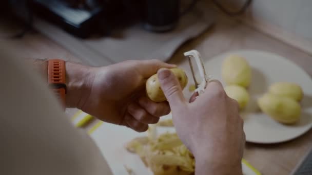 学士男は台所で自宅で夕食を準備します 健康的な食べ物のテーマは 男性はテーブルの上に生のジャガイモのクローズアップで肌を皮を剥きます 農場の非遺伝子組み換え野菜 ジャガイモをむいた男の手 — ストック動画