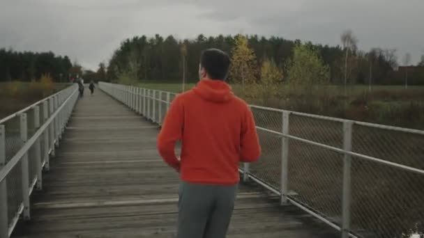 寒い秋の天候で荒野の木製の橋を渡って国立公園を通って走るオレンジ色のパーカーの男性 スポーツマンは田舎の木の歩道橋をジョギングするフィットネスワークアウトを行います — ストック動画
