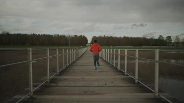 田舎をジョギングするクロスカントリー 国立公園の木製の橋を渡って走るオレンジのスウェットシャツの男 野生動物の歩道の木製の歩道橋の上を走るランナー 寒い秋の天気でトレイルラン — ストック動画