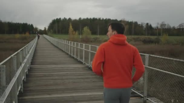 オレンジのスウェットシャツを着た男性ジョガーは 秋にスカンジナビアの自然の中で木の橋を渡って走ります 選手は寒い天候の中 歩道橋を渡って走る 健康的なライフスタイル 国立公園内のトレイルラン — ストック動画