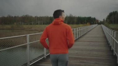 Turuncu kapüşonlu bir erkek soğuk sonbahar havasında ağaçtan köprüye doğru koşuyor. Sporcu, kırsal kesimdeki bir kereste köprüsünde koşuya çıkar.. 