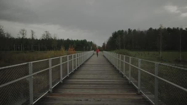 穿着橙色运动衫的慢跑者穿过斯堪的纳维亚半岛自然的木桥 运动员在寒冷的天气里跑过人行天桥 健康的生活方式在国家公园里的小径 — 图库视频影像