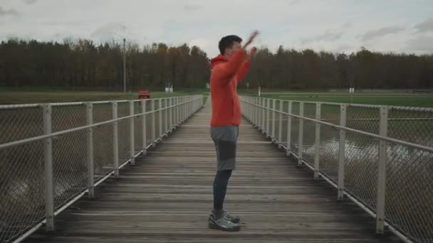 在寒冷的秋天里 穿着橙色和温暖衣服的人在木桥上慢跑前热身 跑步者伸展肌肉 腿和脚 准备跑到户外 跨国跑步 — 图库视频影像