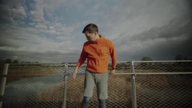 Turuncu ve kalın elbiseli bir adam, soğuk sonbahar havasında ahşap köprüde koşuya çıkmadan önce ısınıyor. Koşucu bacak ve ayakları kasarak dışarı koşmaya hazırlanıyor. Kros koşusu. 