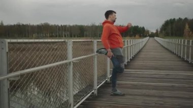 Sporcu açık hava kardiyo çalışmasından önce ısınıyor. Bir koşucu soğuk sonbahar havasında tahta bir köprüde koşmak için kaslarını ısıtır. Sağlıklı bir yaşam tarzı. Kırsal kesim. İz çalışmaları. 