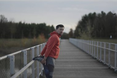 Sporcu açık hava kardiyo çalışmasından önce ısınıyor. Bir koşucu soğuk sonbahar havasında tahta bir köprüde koşmak için kaslarını ısıtır. Sağlıklı bir yaşam tarzı. Kırsal kesim. İz çalışmaları