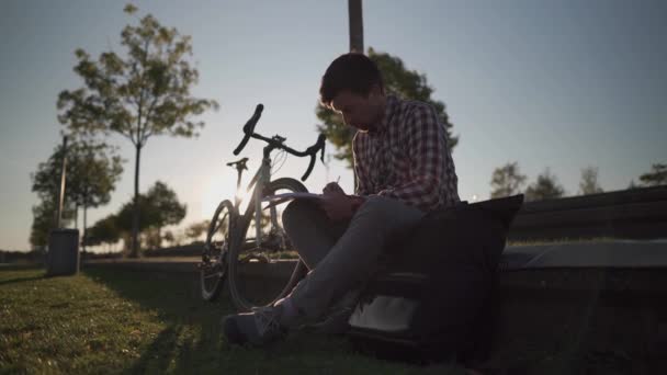 男学生坐在自行车旁边的公园里 在笔记本上记笔记 在公园里学习和做作业的家伙骑自行车来到公园 生活方式 户外教育 大学校园 — 图库视频影像