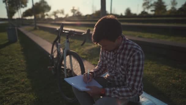 自転車の近くの公園に座ってノートを取っている男性学生 男は自転車で公園に来る彼の宿題を勉強し 公園でやっている ライフスタイル屋外教育 大学キャンパス — ストック動画