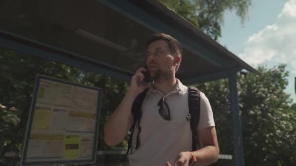 男はバス停でバスを待っている間に腕時計で時間をチェックし 遅れた約束を警告するために呼び出し 男性はドイツでの輸送を遅らせている間に携帯電話で通信します いらいらする男は遅い — ストック動画
