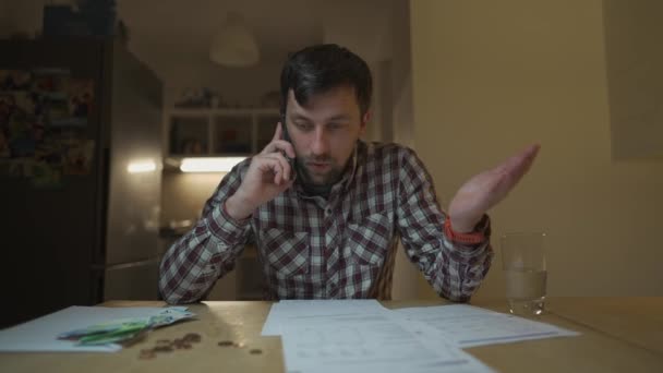 男は台所のテーブルに座っている家で夕方にユーティリティ法案を得た後 電話で主張する 銀行の領収書計算税を見て電話で身に着けている男性 請求書の文書を調べます — ストック動画