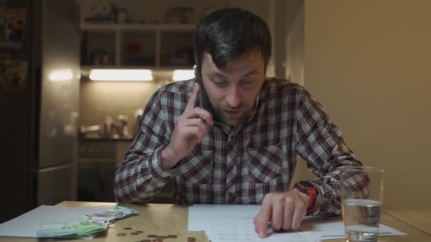 男は台所のテーブルに座っている家で夕方にユーティリティ法案を得た後 電話で主張する 銀行の領収書計算税を見て電話で身に着けている男性 請求書の文書を調べます — ストック動画