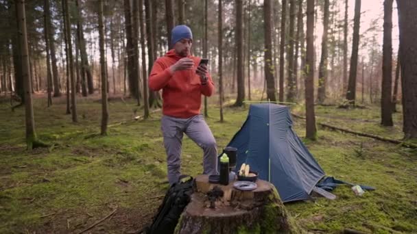男子徒步旅行者在秋天的树林里徒步旅行时 在帐篷营地用智能手机喝茶和上网 背包客通过电话在地图上标明路线 并在帐篷后边喝一杯热饮 — 图库视频影像