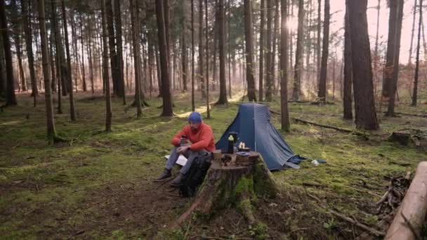 早上在秋天森林的露营地 Tursit坐在帐篷附近的远足垫子上 喝着杯子里的饮料 一边用智能手机浏览互联网 人和健康的生活方式 — 图库视频影像