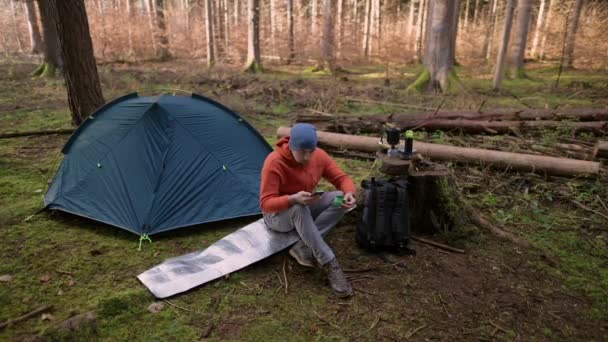 かわいい男ハイカーはテントの近くの旅行マットに座って カップで熱い飲み物を飲みます 秋の森でのバックパッキング旅行で朝食をしながら スマートフォンを使用して男性旅行者 キャンプ場のコンセプト — ストック動画