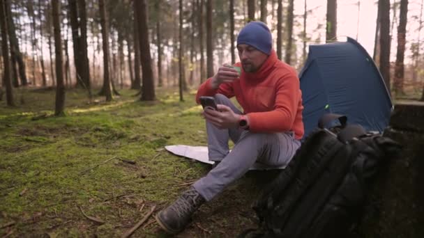 早上在秋天森林的露营地 Tursit坐在帐篷附近的远足垫子上 喝着杯子里的饮料 一边用智能手机浏览互联网 人和健康的生活方式 — 图库视频影像