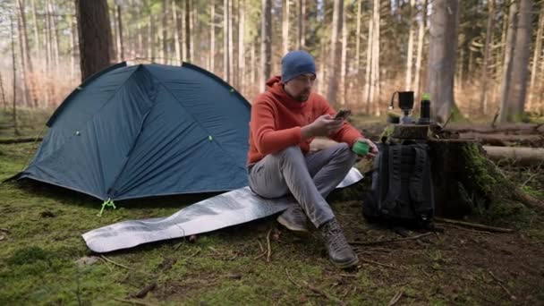 一个可爱的远足男子坐在帐篷附近的旅行垫子上 喝着一杯热饮 在秋天的森林里 在一次背包旅行中 男性旅行者一边吃早饭一边用智能手机 营地概念 — 图库视频影像