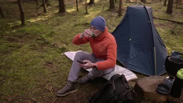 男子徒步旅行者在秋天的树林里徒步旅行时 在帐篷营地用智能手机喝茶和上网 背包客通过电话在地图上标明路线 并在帐篷后边喝一杯热饮 — 图库视频影像