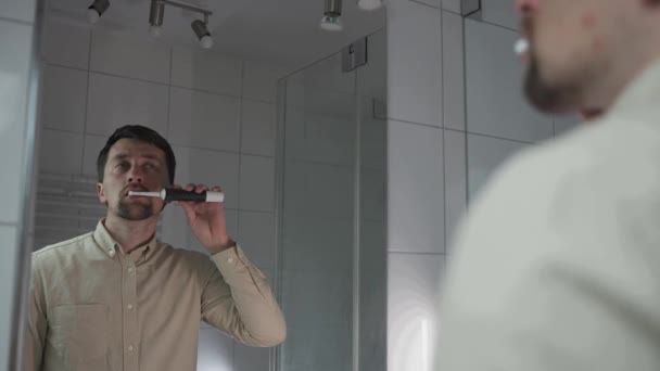 白人在浴室里用电动牙刷刷牙 透过镜子看到自己的倒影 早上的卫生习惯口服灌洗器 早上的例行公事美丽的微笑 — 图库视频影像