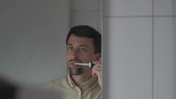 仕事の前に朝のバスルームで電動歯ブラシでシャツのクリーニング歯の男 鏡を通して反射ビュー 電気音響歯ブラシを使う男性 毎日の歯科衛生と口腔衛生 — ストック動画