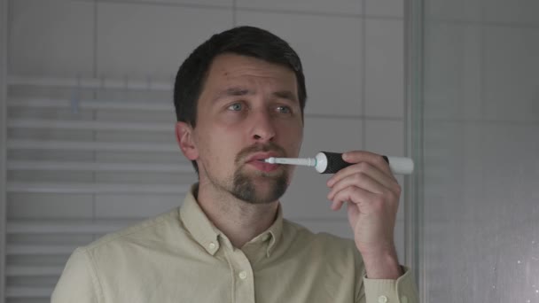 男はシャツで働く前に 午前中に音波充電式電動歯ブラシで口を磨く 鏡の中の反射を通して見る 毎日のボディケア 毎日の歯科衛生と口腔衛生 — ストック動画