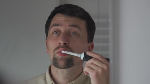 白人男性は 鏡を通して反射のビューは バスルームに電動歯ブラシで彼の歯を磨く 朝の衛生手順 オーラルインジェクター 美しい笑顔のための朝のルーチン — ストック動画