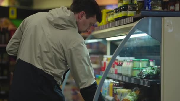男人选择沙拉时 手里拿着一包牛奶 穿着夹克在超市里 人们买日用品 顾客在商店里挑选准备好的盒装沙拉 健康饮食 那家伙买了一盒生菜 — 图库视频影像