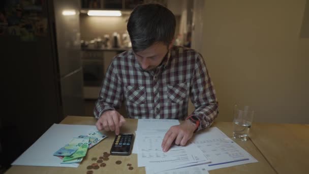 不満を持つ男は請求書を見て 台所のアパートで計算機でお金を計算します 男性の金融をチェックする落ち込んでいる 現金問題だ 請求書と税金の支出を数える 公共サービスの請求 — ストック動画