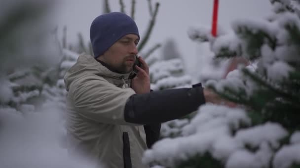 新年の祭典のシンボル 電話ショッピングを持つ男性と雪の日の外の冬に市場で選択します 休日の買い物 電話で話すとモミの木を選択するためのアドバイスを求める — ストック動画