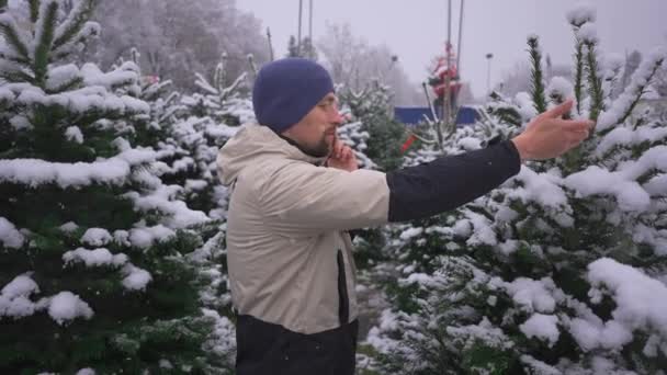 男はクリスマスマーケットでクリスマスツリーを選択し 雪の日に彼の家族から電話で正しい選択にヒントを取ります 地元のファーマーズマーケットでモミの木を拾い 携帯電話で話す — ストック動画