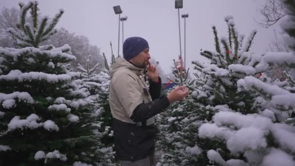新年庆祝活动的象征 有电话购物的男人 冬天在外面的大雪中挑选 假日购物 电话交谈 询问有关选择冷杉树的建议 — 图库视频影像