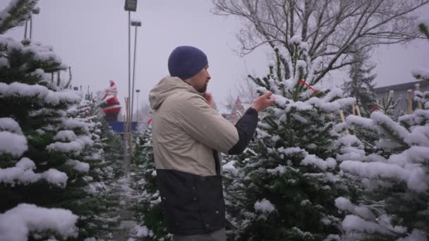 男人在雪地的户外市场上挑选圣诞树 与妻子通电话 并建议她做出正确的选择 圣诞节和新年假期的象征 纽约和圣诞节的假日购物 — 图库视频影像