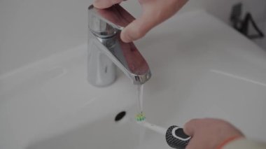 Erkek elinin banyodaki musluğun altında elektrikli diş fırçası tutuşu. Adam dişlerini fırçaladıktan sonra ultrasonik diş fırçasını musluk suyuyla yıkıyor. Ağız temizliği kavramının ayrıntıları