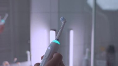 Erkek elinin beyazlı ultrasonik diş fırçasını açıp kapayarak banyonun arka planında mavi bir arka ışık oluşturduğu yakın plan. Ağız bakımı ve beyaz dişler. Elektrikli diş fırçası. Diş temizleyici güç aracı. 