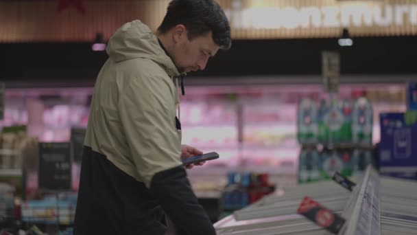 スーパーマーケットの冷凍庫から冷凍食品を選択してスマートフォンを持つ男 男性は市場の冷蔵庫に立っている間 電話で製品情報を読み取ります スマートフォンのショッピングリスト 冷凍製品 — ストック動画