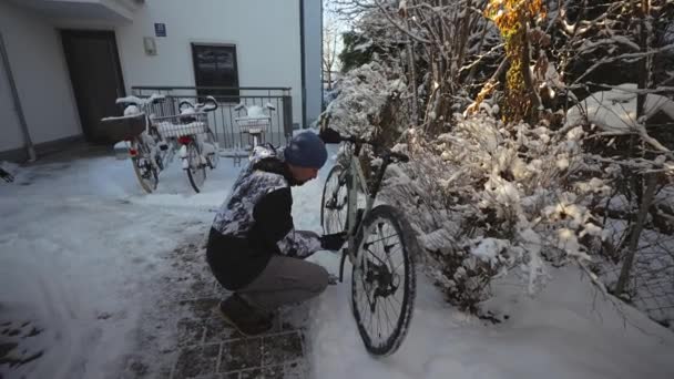 男サイクリストは駐車場にそれを取る前に冬に裏庭で彼の自転車を磨く 冬のサイクリングや自転車のクリーニング 家の外の自転車の車輪から男性のスクレープ雪 — ストック動画