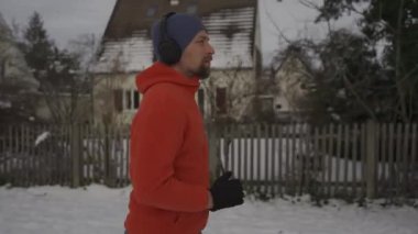 Man jogging on snowy path through park in city listening to music in earphones in winter in Germany. Athletic man working out wearing headphones in freezing cold weather. Running in snow in winter. 