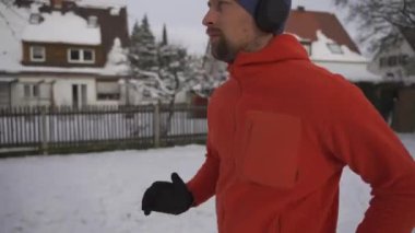 Man jogging on snowy path through park in city listening to music in earphones in winter in Germany. Athletic man working out wearing headphones in freezing cold weather. Running in snow in winter. 