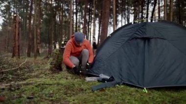 Tourist makes camp in woods, sets up tent in meadow in fall forest, prepares for an overnight stay in campsite. Backpacker builds tent on solo autumn trip in forest. Hiking and staying in nature. 