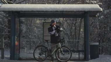 Frozen male cyclist waiting for bus at halt stop with bicycle in winter in snowy weather. Velocipede broke down and man waits long time for transport at the bus stop in winter after a snow storm. 