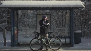 Cyclist extremely cold to ride bicycle in heavy frost and snow in winter and waiting for iced bus at stop, jumping and rubbing his hands. Snowy weather in Germany. A froze commuter on a bus stand. 